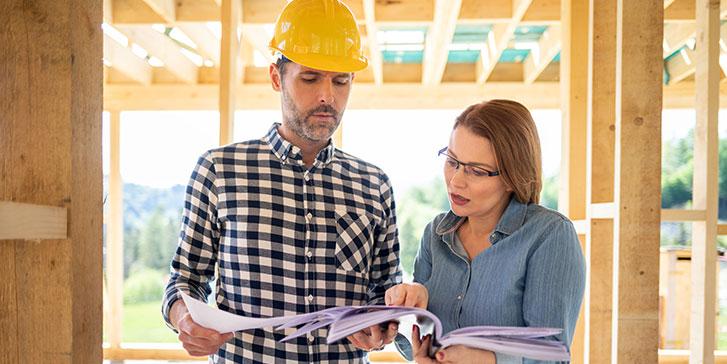 A remodeling contractor in a yellow hard hat and a plaid shirt stands next to a woman in glasses and a blue shirt. They are looking at and discussing papers together at a construction site with a wooden frame structure.
