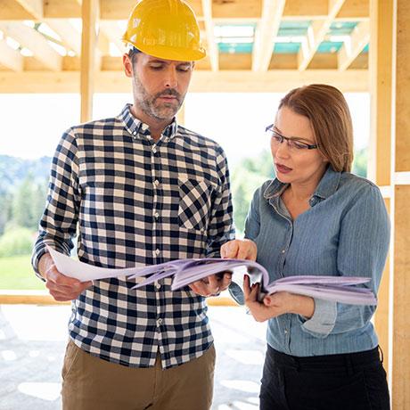 A remodeling contractor in a yellow hard hat and a plaid shirt stands next to a woman in glasses and a blue shirt. They are looking at and discussing papers together at a construction site with a wooden frame structure.