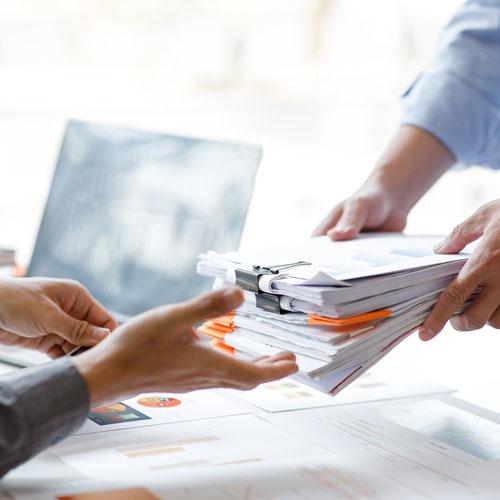 Two people exchanging a large stack of documents with paper clips and folders in an office setting during the process of business formation; one person is handing the papers over, while the other reaches out to receive them. A laptop is visible in the background.