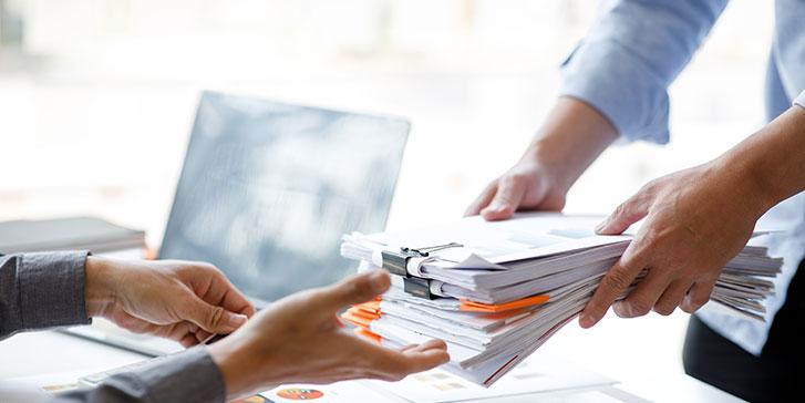 Two people exchanging a large stack of documents with paper clips and folders in an office setting during the process of business formation; one person is handing the papers over, while the other reaches out to receive them. A laptop is visible in the background.
