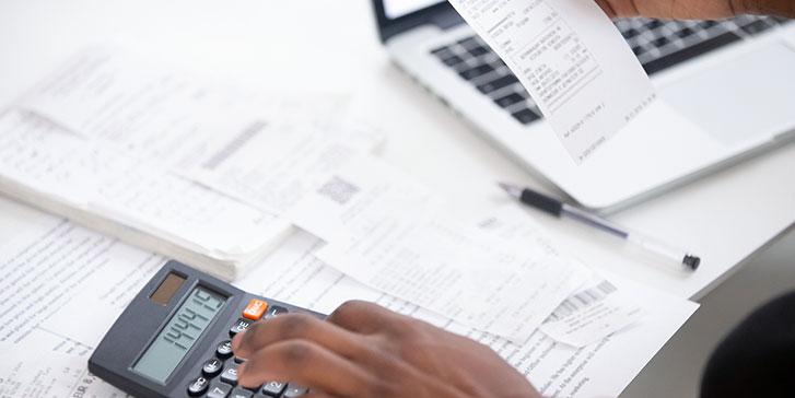 A person uses a calculator while holding a receipt, with more receipts, papers, a pen, and a laptop visible on the desk, suggesting they are doing financial calculations to determine their sales tax liability.