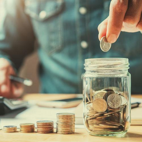 A person drops a coin into a glass jar filled with coins, while using a calculator and holding a pen in the background. Stacks of coins and paperwork are also visible on the table, representing claiming tax incentives