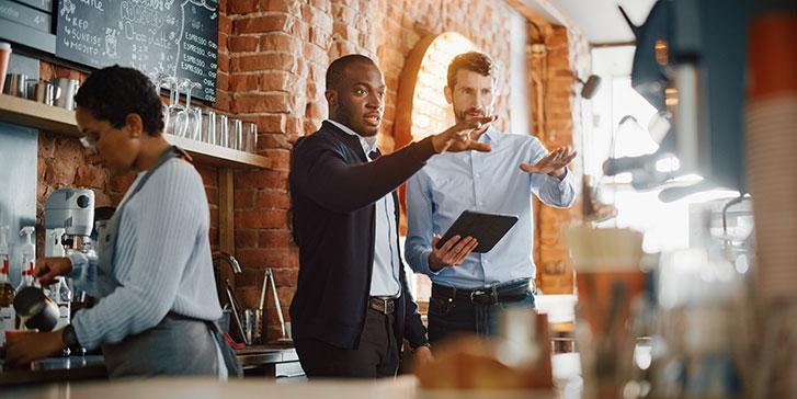 Two men discuss the unique situation of taxes in the hospitality industry while standing behind the counter of a coffee shop; one holds a tablet. A woman in the foreground prepares a drink behind the counter. The setting is warm with exposed brick walls and a menu on the wall.
