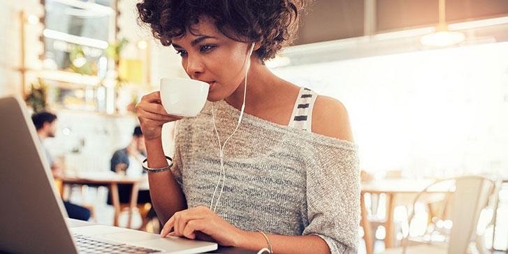 A freelancer with curly hair sits at a table in a café, wearing earphones and drinking from a white cup while using a laptop. An empty saucer is on the table, and other people are in the background. The freelancer is researching information about her tax liability as an independent contractor