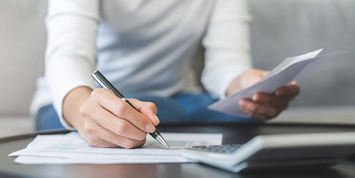 A person wearing a white long-sleeve shirt sits at a table, holding a paper in one hand and writing with a pen on tax documents with the other. A calculator is partially visible on the table.