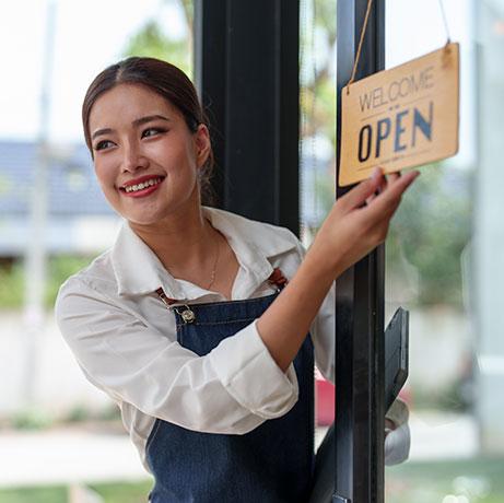 A smiling small business owner wearing an apron opens a glass door and flips a sign to Welcome, We Are Open, greeting customers as she stands inside a shop or café.