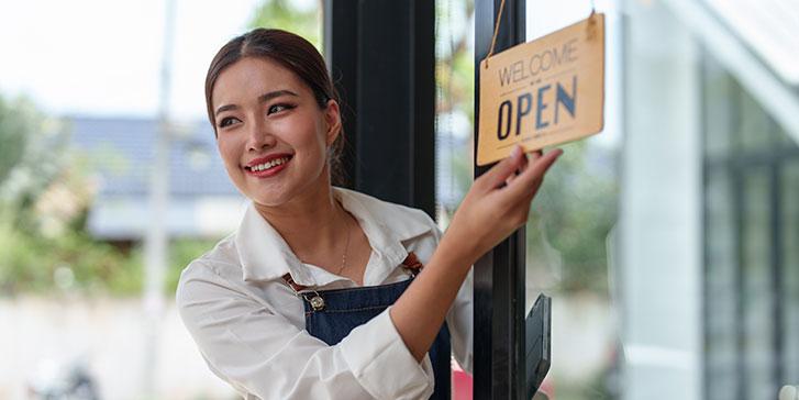 A smiling small business owner wearing an apron opens a glass door and flips a sign to Welcome, We Are Open, greeting customers as she stands inside a shop or café.