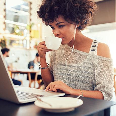 A freelancer with curly hair sits at a table in a café, wearing earphones and drinking from a white cup while using a laptop. An empty saucer is on the table, and other people are in the background. The freelancer is researching information about her tax liability as an independent contractor